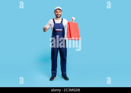 Full length happy professional courier in uniform pointing at shopping bags and smiling to camera, delivering parcel with goods ordered online at fash Stock Photo