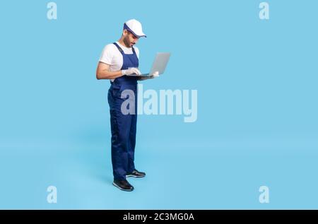 Full length professional workman in uniform and protective gloves typing on laptop keyboard, having online communication with client, reading order fo Stock Photo