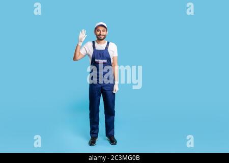 Full length friendly handyman in overalls and protective gloves standing, waving hello, welcoming with hi gesture. Profession of service industry, hou Stock Photo