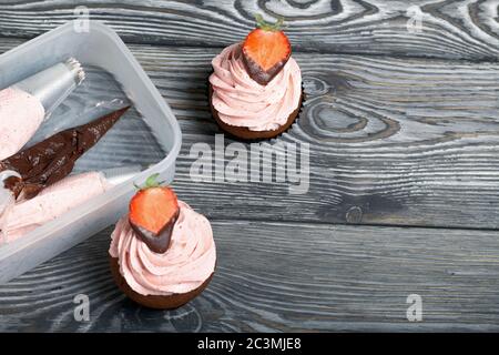 Chocolate cupcakes with strawberry cream. Decorated with half strawberries in chocolate. Next to the blanks and ingredients for cupcakes. Stock Photo