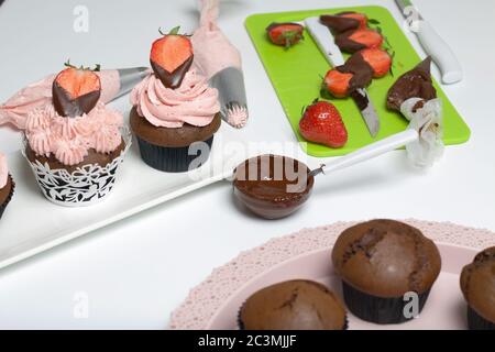 Chocolate cupcakes with strawberry cream. Decorated with half strawberries in chocolate. Next to the blanks and ingredients for cupcakes. Stock Photo