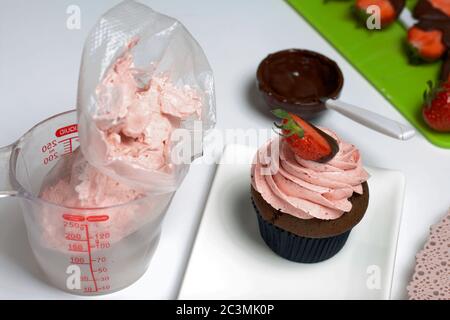 Chocolate cupcakes with strawberry cream. Decorated with half strawberries in chocolate. Next to the blanks and ingredients for cupcakes. Stock Photo