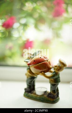 Aroma lamp with two cooks carrying food in a fruit pot. Kitchen utensils on a windowsill with roses outside the window Stock Photo