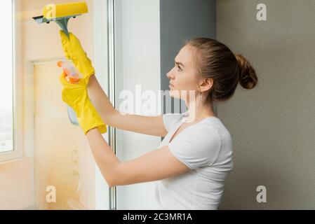Cute adult girl washes a window with a scraper. Spring cleaning. An employee of a cleaning company wipes plastic windows in the house. Stock Photo