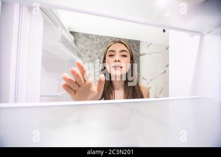 Young woman on kitchen during quarantine. Look at empty fridge shelves with no food on it. Hungry and can't cook. Back view of woman doesn't know what Stock Photo