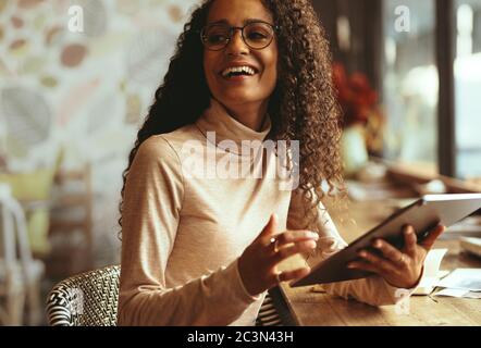Smiling woman sitting at a cafe with her digital tablet. Female with curly hair looking away and smiling while sitting at a coffee shop table. Stock Photo