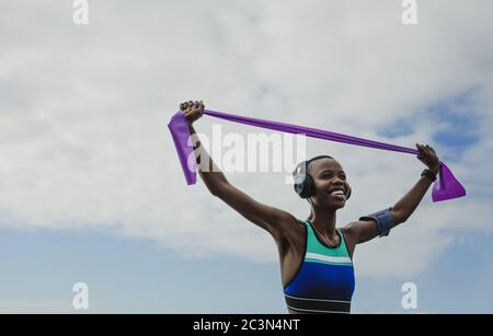 Happy woman in sportswear exercises with resistance band. Fitness female wearing headphones doing stretching workout against sky. Stock Photo