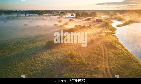Summer misty sunrise on meadow. Country road on green fields riverbank. Sunlight on large oak trees grove in morning. Aerial view, Belarus, Berezina r Stock Photo