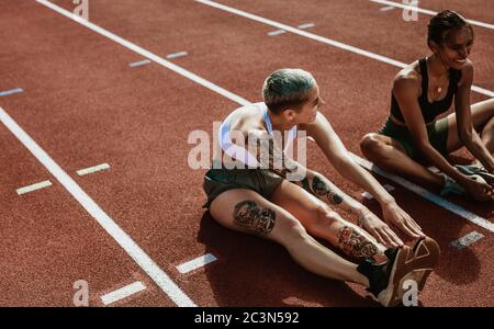 Two female athletes doing stretching exercises sitting on running track. Woman runners sitting on running track stretching legs. Stock Photo