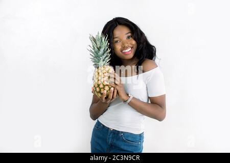 Cheerful attractive african american young woman in jeans and white t-shirt standing and holding pineapple over white background. Beautiful African Stock Photo