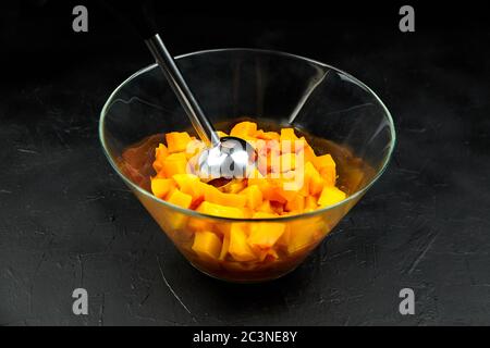 Pumpkin pieces and blender in a glass bowl on black table. Chopped butternut squash in salad dish. Cooking vegetable food, pumpkin puree (mash) recipe Stock Photo