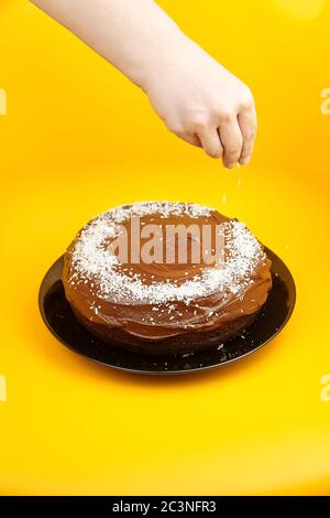 Chocolate cake decorated with flaked coconut, homemade pie on yellow surface. Femake hand scattering coconut flakes. Home cake with cocoa ingredient o Stock Photo