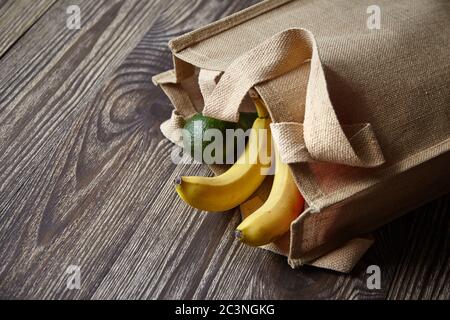Reusable shopping bag with fresh fruits. Natural eco friendly material. Yellow bananas, green avocado, hessian or jute sack on brown wooden table. Use Stock Photo