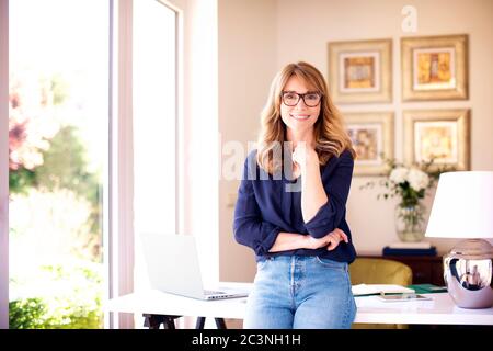 Portrait shot of smiling mature woman standing at desk at home while looking at camera and smiling. Stock Photo