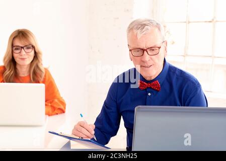 Shot of senior businessman wearing bow tie and glasses while using laptop in the office. Middle aged businesswoman sitting behind him and working on n Stock Photo