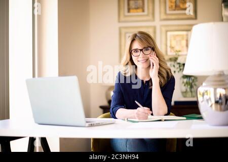 Shot of smiling mature woman making a phone call and using laptop while working online. Home office. Stock Photo