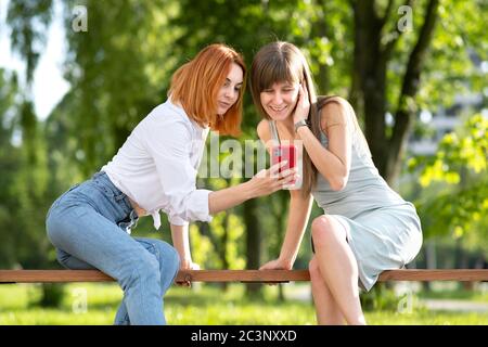 Two young female friends sitting on a bench in summer park taking a selfie by smartphone camera. Stock Photo