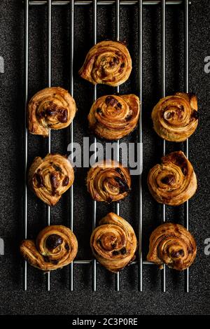 Puff pastry snails with custard and raisins cooling down on a steel grate, deep grey background, shot from above Stock Photo