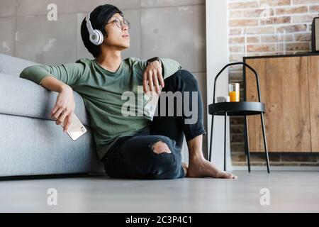 Image of handsome young asian man wearing headphones holding cellphone while resting in apartment Stock Photo