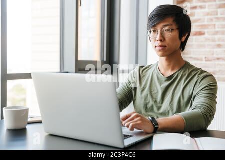 Image of handsome young asian man wearing eyeglasses using laptop while sitting at table in apartment Stock Photo