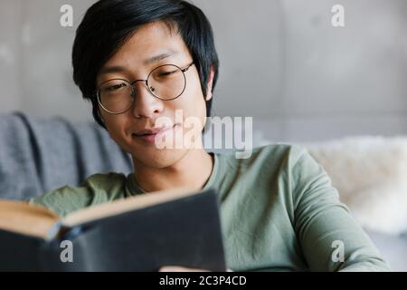 Image of handsome young asian man wearing eyeglasses reading book while sitting on couch in apartment Stock Photo