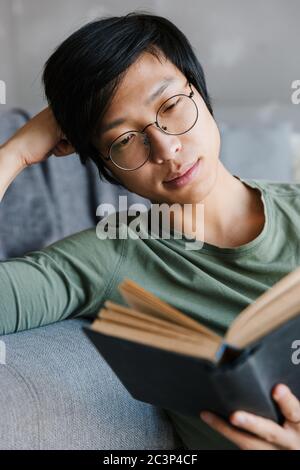 Image of handsome young asian man wearing eyeglasses reading book while sitting on couch in apartment Stock Photo