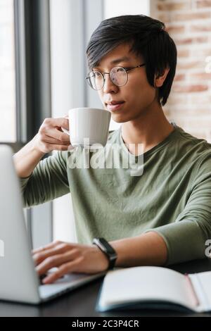 Image of handsome young asian man wearing eyeglasses holding tea cup while using laptop in apartment Stock Photo