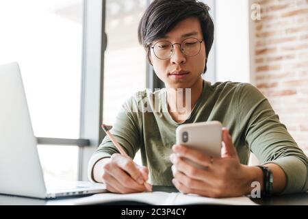 Image of handsome young asian man wearing eyeglasses using laptop and cellphone in apartment Stock Photo