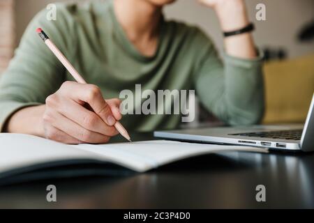 Image of young asian man wearing eyeglasses writing down notes and using laptop in apartment Stock Photo