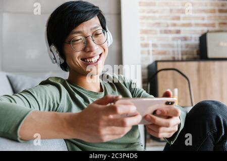 Image of handsome young asian man wearing headphones using cellphone while resting in apartment Stock Photo