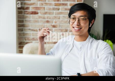 Image of handsome young asian man wearing earphones using laptop while sitting at table in office Stock Photo