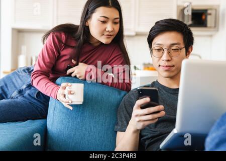 Photo of focused asian couple using smartphone and laptop while drinking coffee on sofa at home Stock Photo