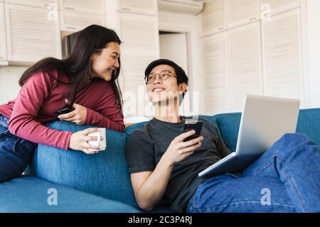 Photo of smiling asian couple using smartphone and laptop while drinking coffee on sofa at home Stock Photo