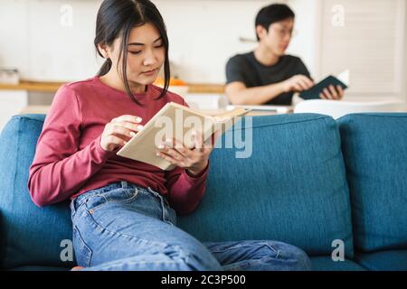 Photo of young concentrated asian couple reading books while sitting at cozy room Stock Photo