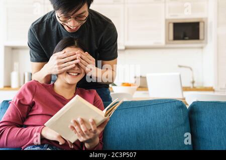 Photo of joyful asian man covering girlfriend's eyes while she reading book on sofa in cozy room Stock Photo