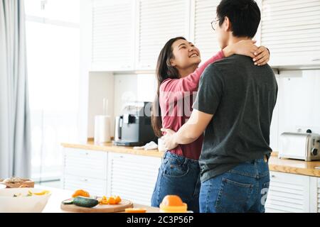 Beautiful happy young asian couple embracing while standing in the kitchen Stock Photo