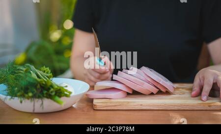 Young woman cutting sausage on wooden board outdoors. Women's hands cutting sausage goods with knife on chopping board. Stock Photo