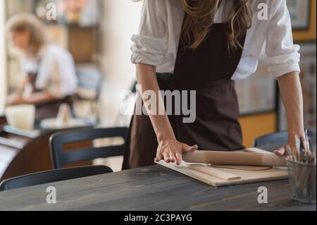 Female hands flatting piece of clay lying on fabric using wooden rolling pin in ceramic studio. Stock Photo