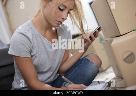 Young blonde female writing in her notepad on the floor surrounded by unpacked boxes Stock Photo