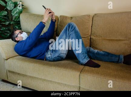 Caucasian man wearing a medical mask lying on a sofa doing a selfie Stock Photo