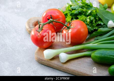 Old wooden cutting board with fresh vegetables on stone concrete table. Close-up view on salad ingredients: tomatoes, cucumbers, onion, parsley Stock Photo