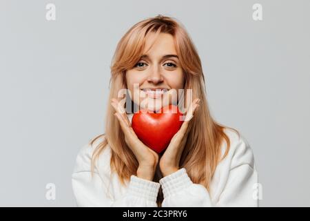 Portrait of joyful attractive woman with strawberry blonde hair hold red heart (Valentine day symbol),looking at camera, closeup, isolated on light ba Stock Photo