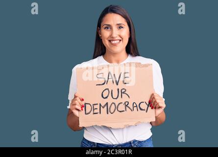 Young beautiful woman holding save our democracy cardboard banner looking positive and happy standing and smiling with a confident smile showing teeth Stock Photo