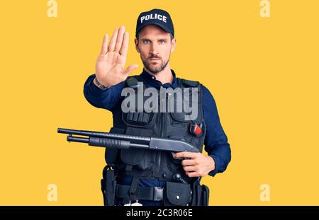 Young handsome man wearing police uniform holding shotgun stressed and frustrated with hand on head, surprised and angry face Stock Photo