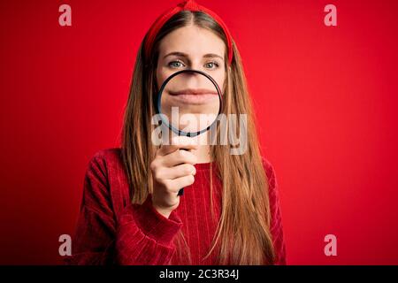 Young beautiful redhead detective woman using magnifying glass over isolated red background with a confident expression on smart face thinking serious Stock Photo
