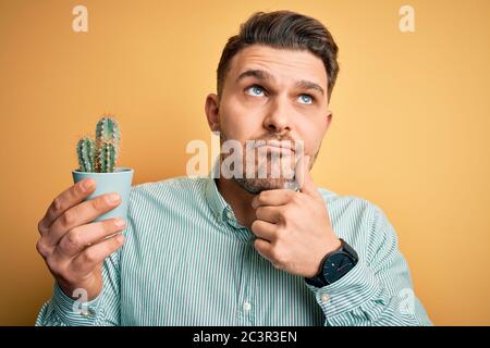 Young man with blue eyes holding fresh cactus plant pot over yellow background serious face thinking about question, very confused idea Stock Photo