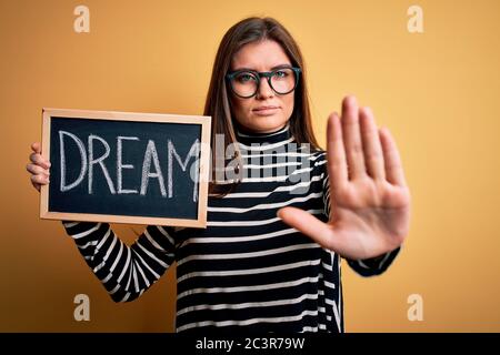 Young beautiful woman with blue eyes holding blackboard with dream message word with open hand doing stop sign with serious and confident expression, Stock Photo
