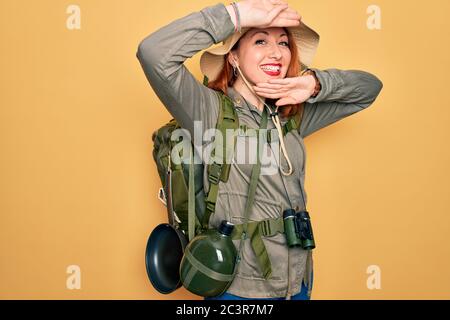 Young redhead backpacker woman hiking wearing backpack and hat over yellow background Smiling cheerful playing peek a boo with hands showing face. Sur Stock Photo