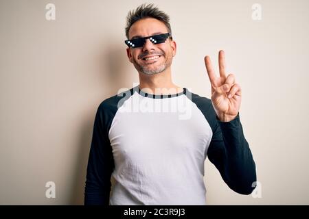 Young handsome man wearing funny thug life sunglasses meme over white background smiling with happy face winking at the camera doing victory sign. Num Stock Photo