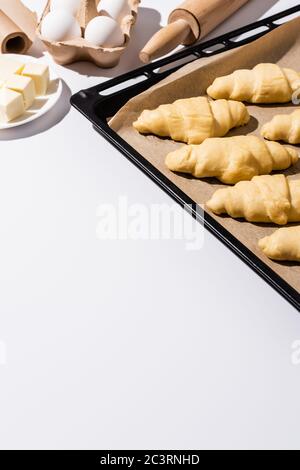 selective focus of raw croissants on baking tray near rolling pin, parchment paper, butter, eggs on white background Stock Photo